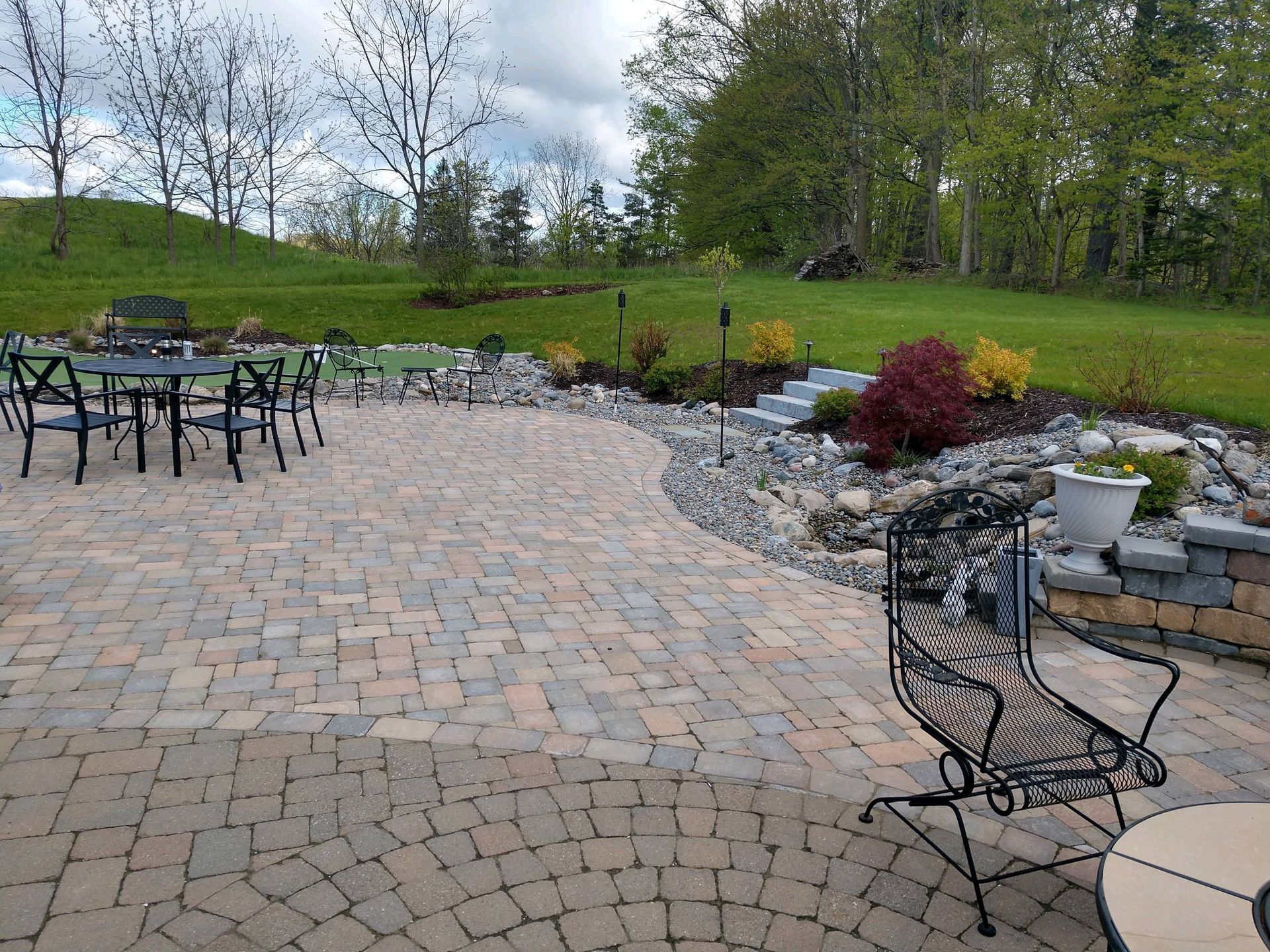 Brick patio with metal furniture, bordering a rock garden and grassy hill with trees.
