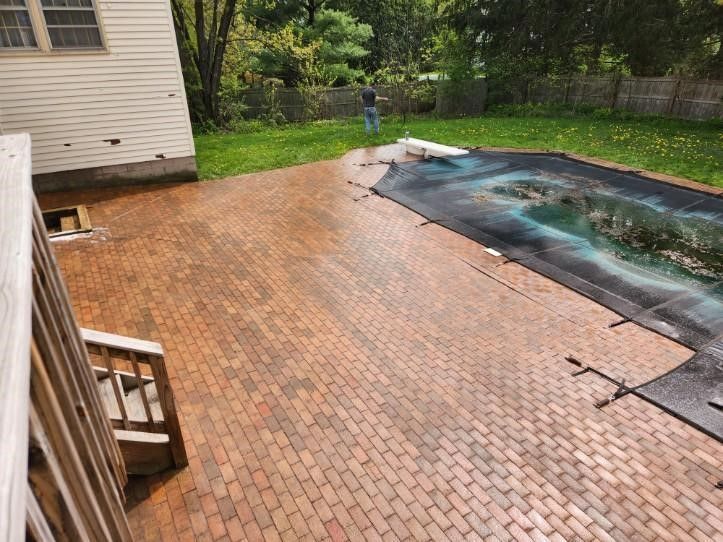 Brick patio next to a pool with a black cover; person in the background, green grass, and white house.