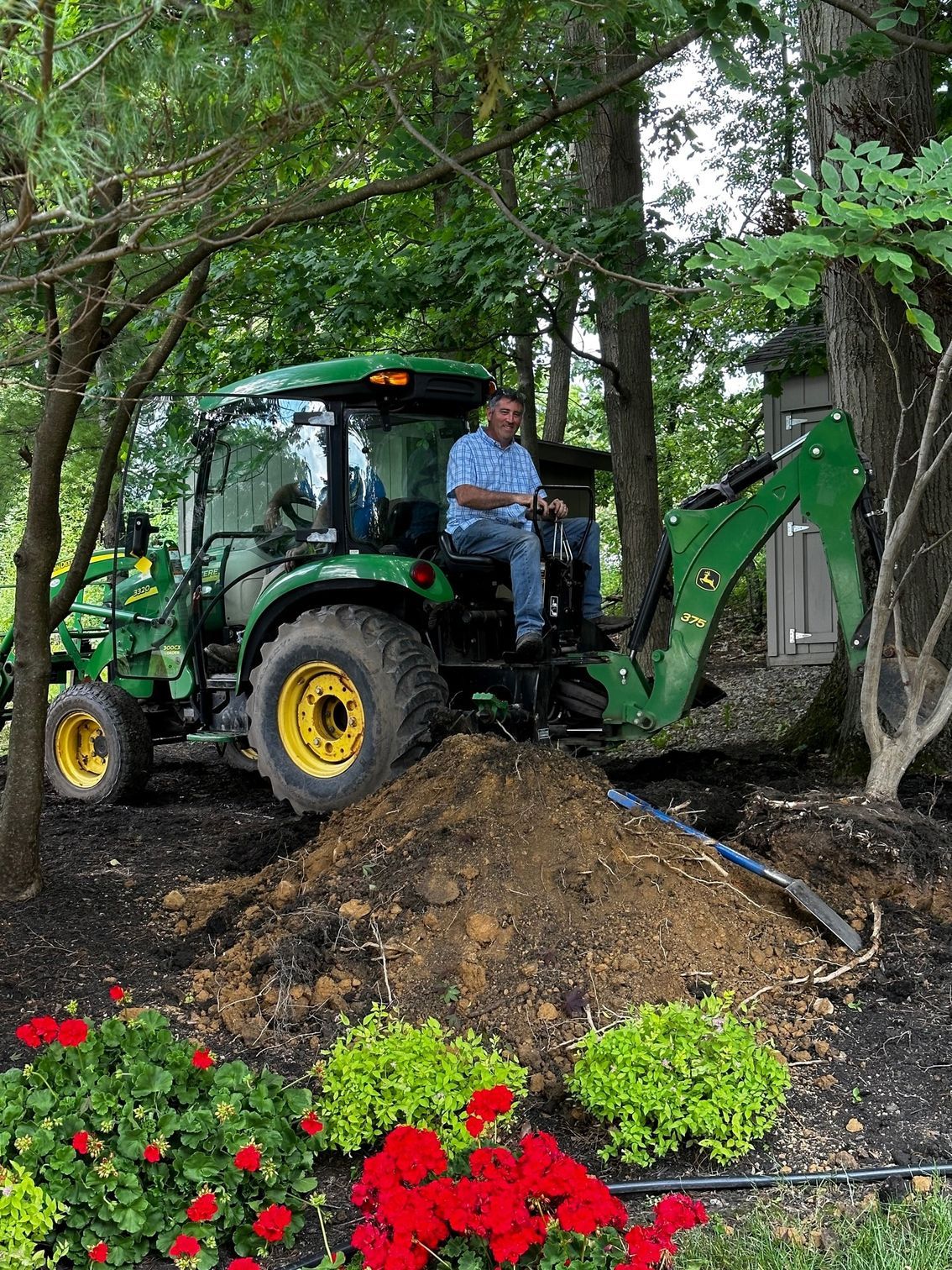 Man operating a green John Deere tractor with a backhoe, digging in a garden bed with red flowers.