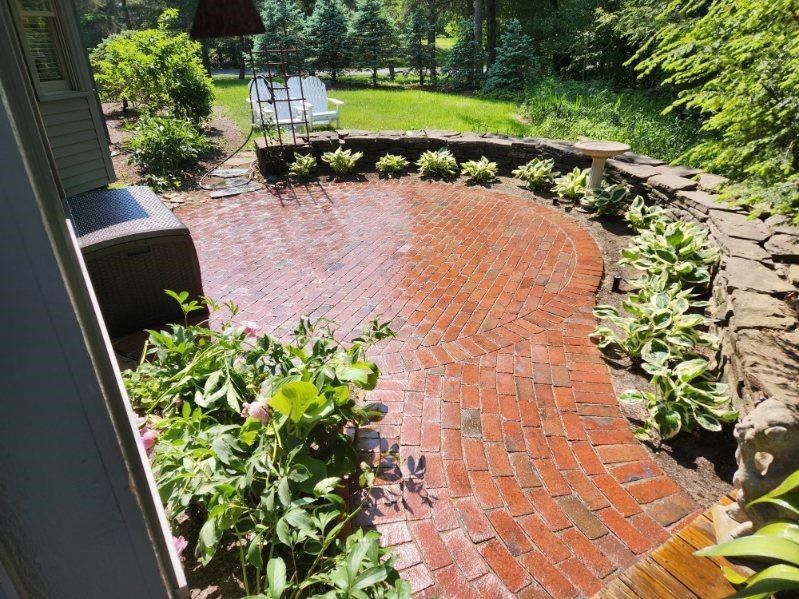 Brick patio with curved path, bordered by stone wall and plants, leading to a lawn with white chairs.