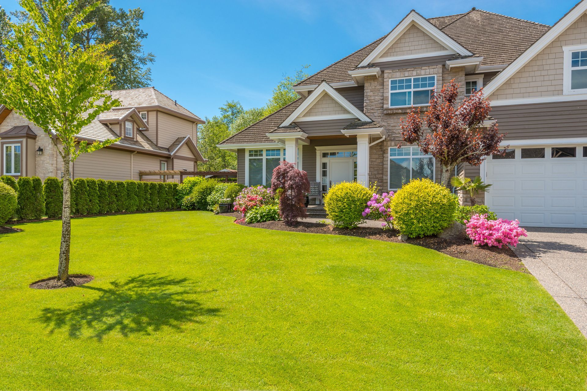 Green lawn in front of a two-story house with a driveway. Landscaping includes trees, bushes, and colorful flowers.