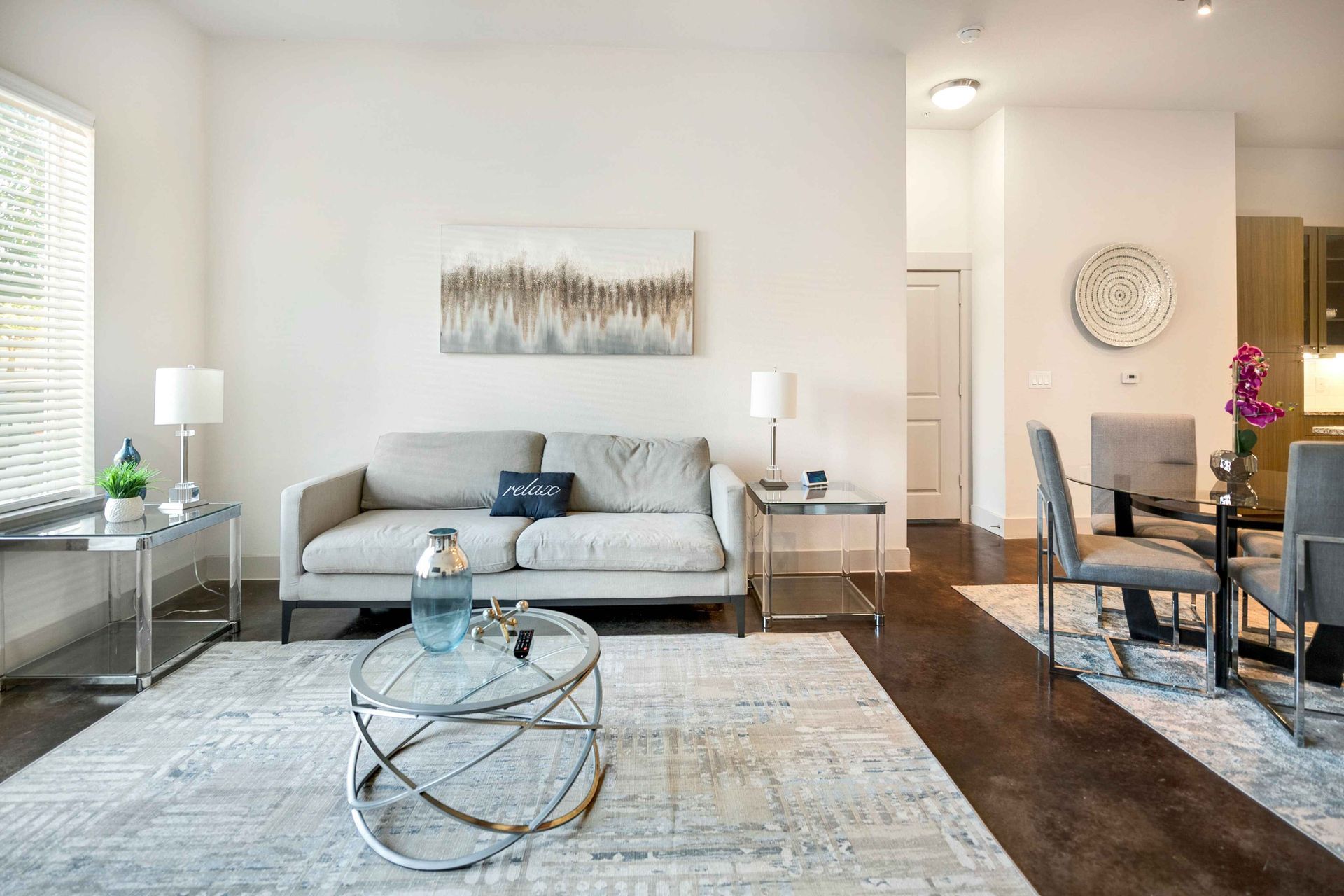 Living room with gray sofa, silver tables, area rug, and dining table. White walls, wood floor.