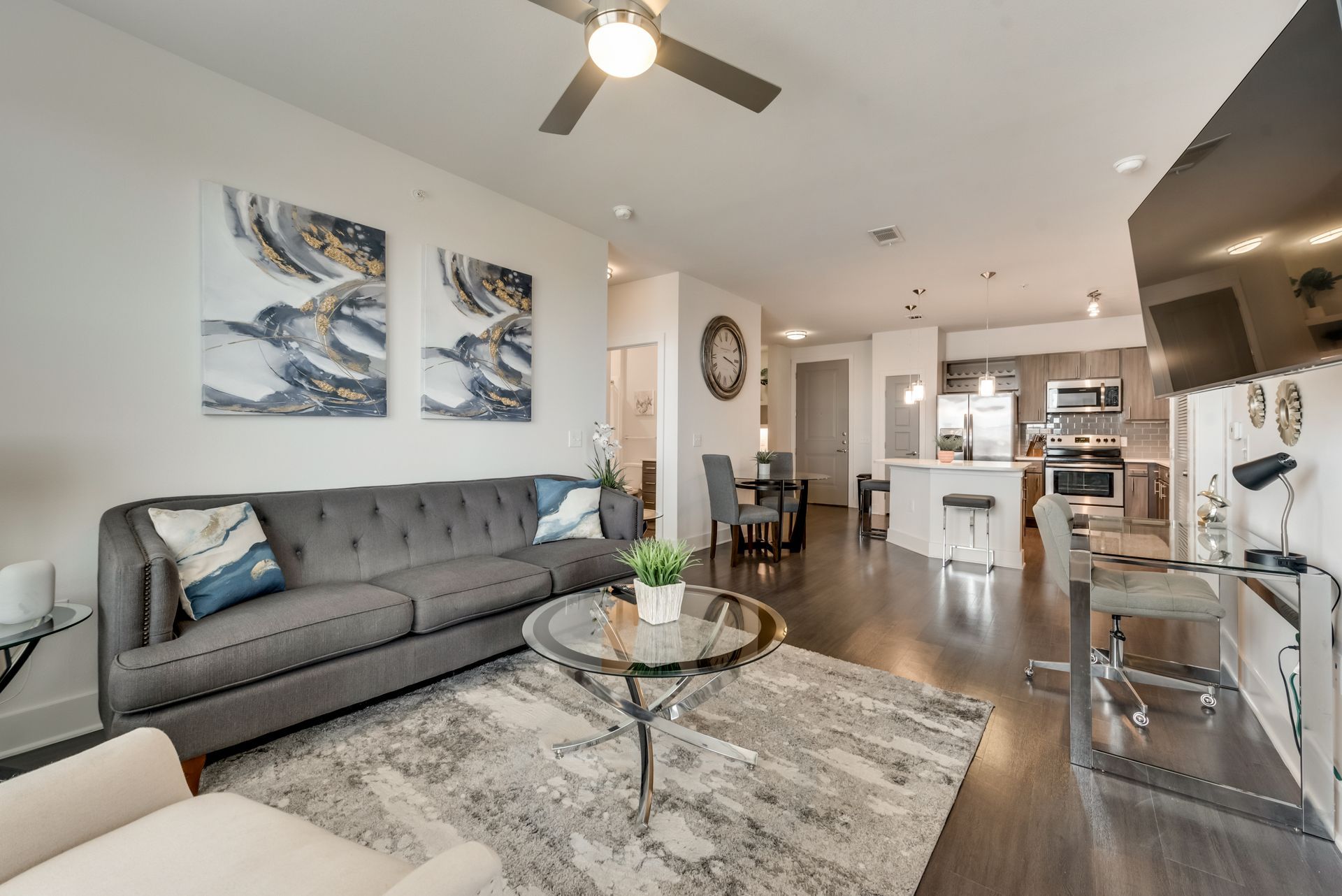 Modern apartment living room with gray sofa, abstract art, open kitchen, and dark wood floors.
