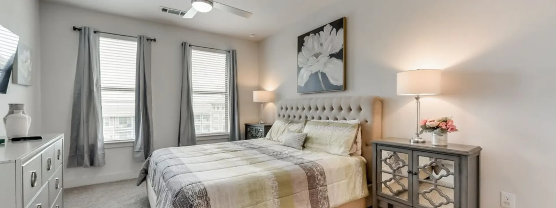 Bedroom with bed, nightstands, windows, and artwork. Gray and neutral colors.