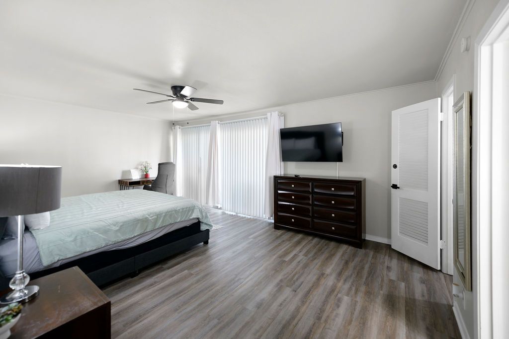 Bedroom with bed, dresser, TV, and sliding doors to balcony. White walls, gray wood floor.