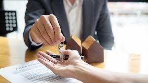 Person in a suit handing keys to another person over a document and wooden house models.