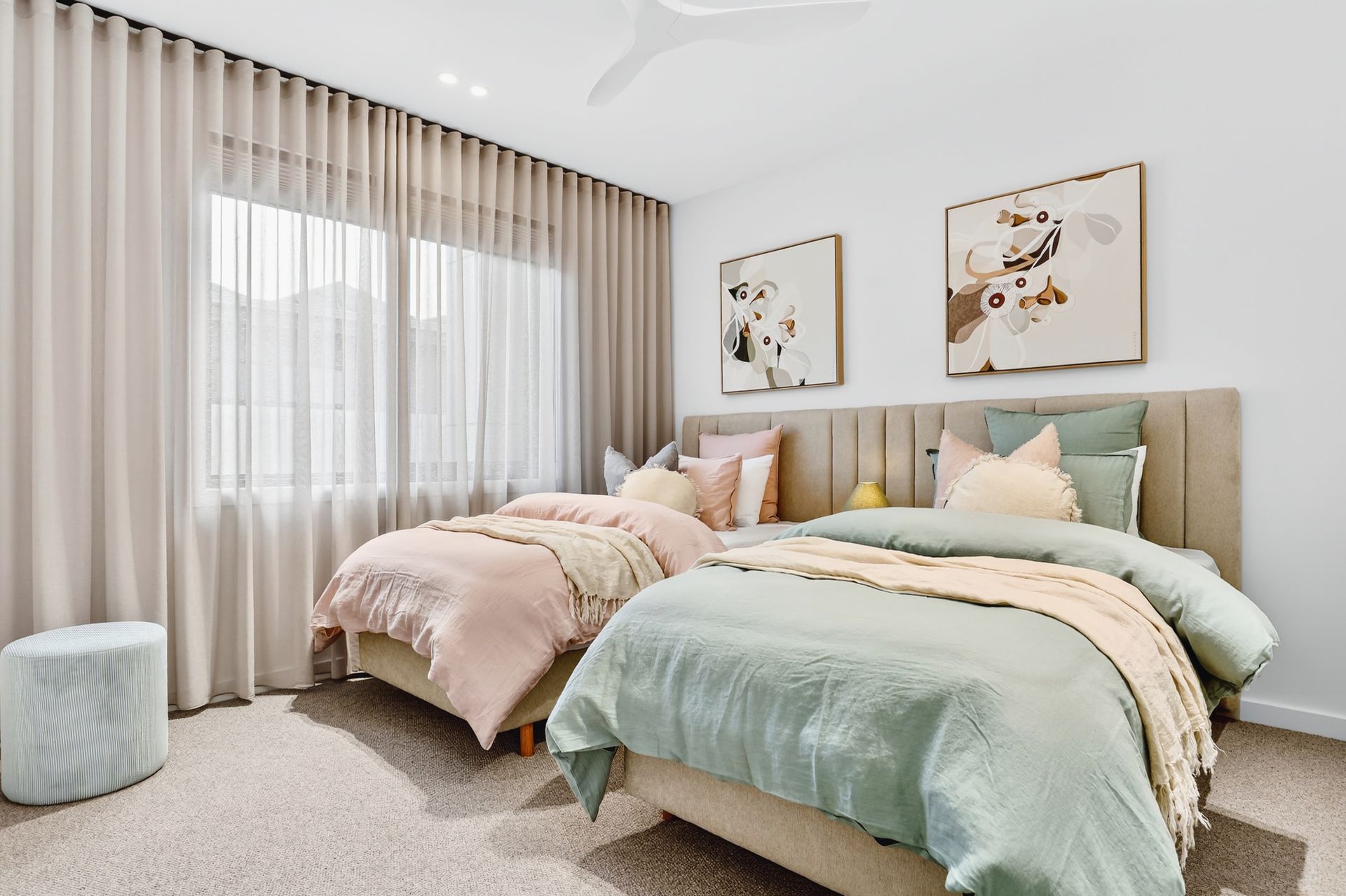Bedroom with two twin beds, pink and green bedding, neutral curtains, art, and a small stool.