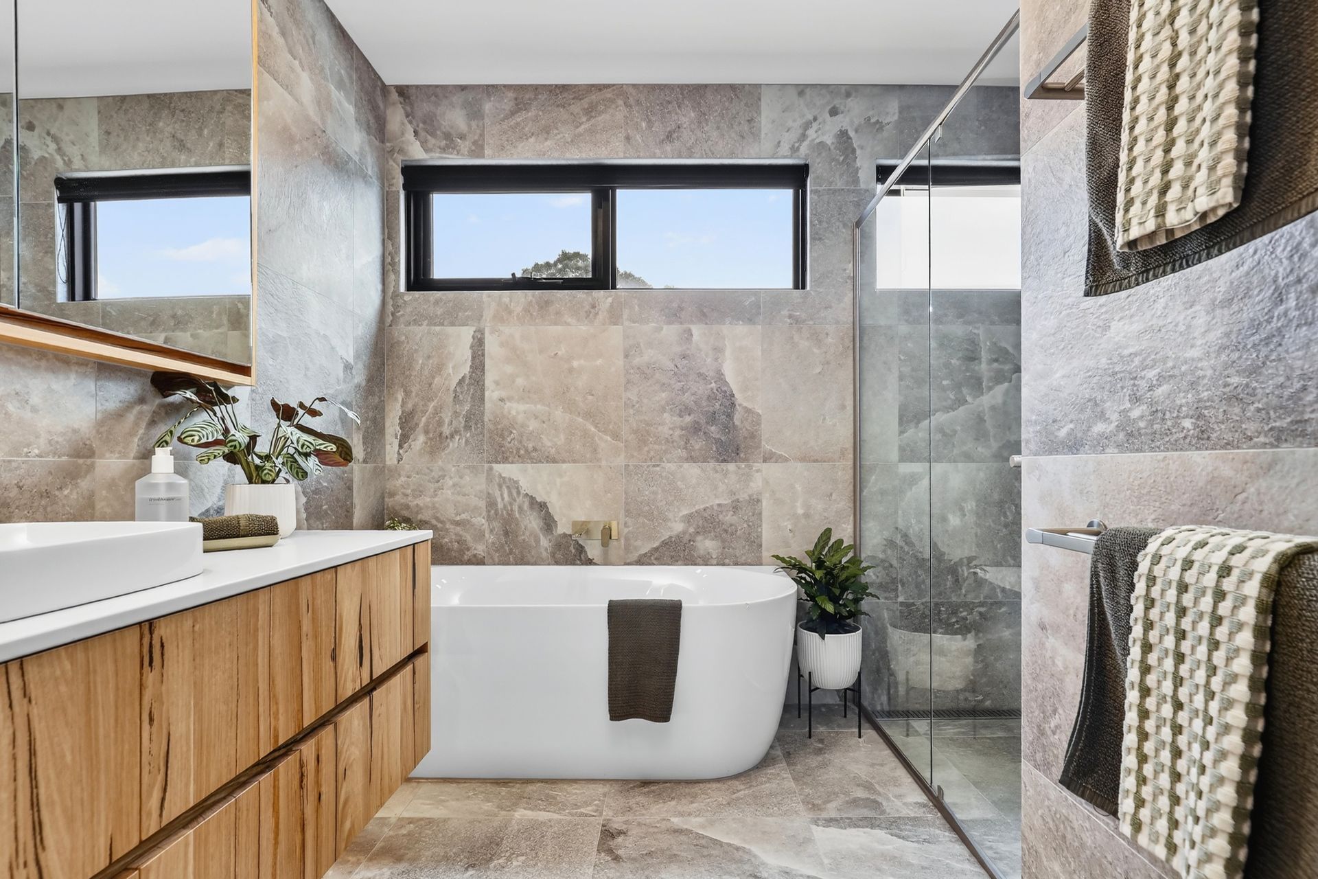 Modern bathroom with stone tile walls, wood vanity, white tub, and glass shower.