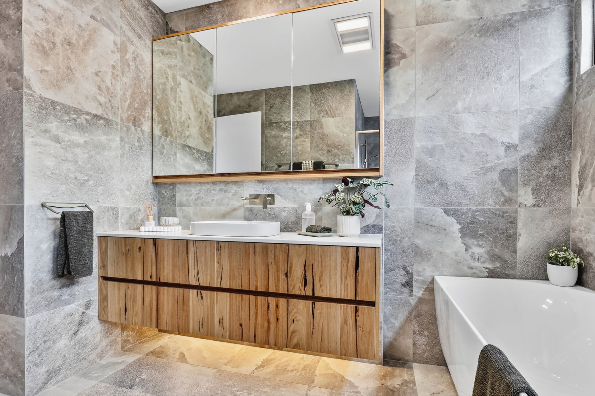 Modern bathroom with wooden vanity, large mirror, stone tiles, and a bathtub.