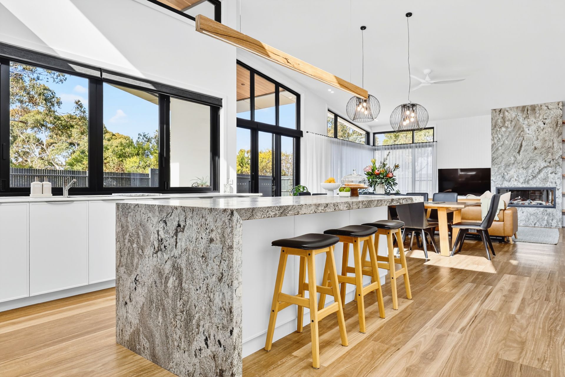 Modern kitchen with granite island, wooden stools, large windows, and open to a dining area.