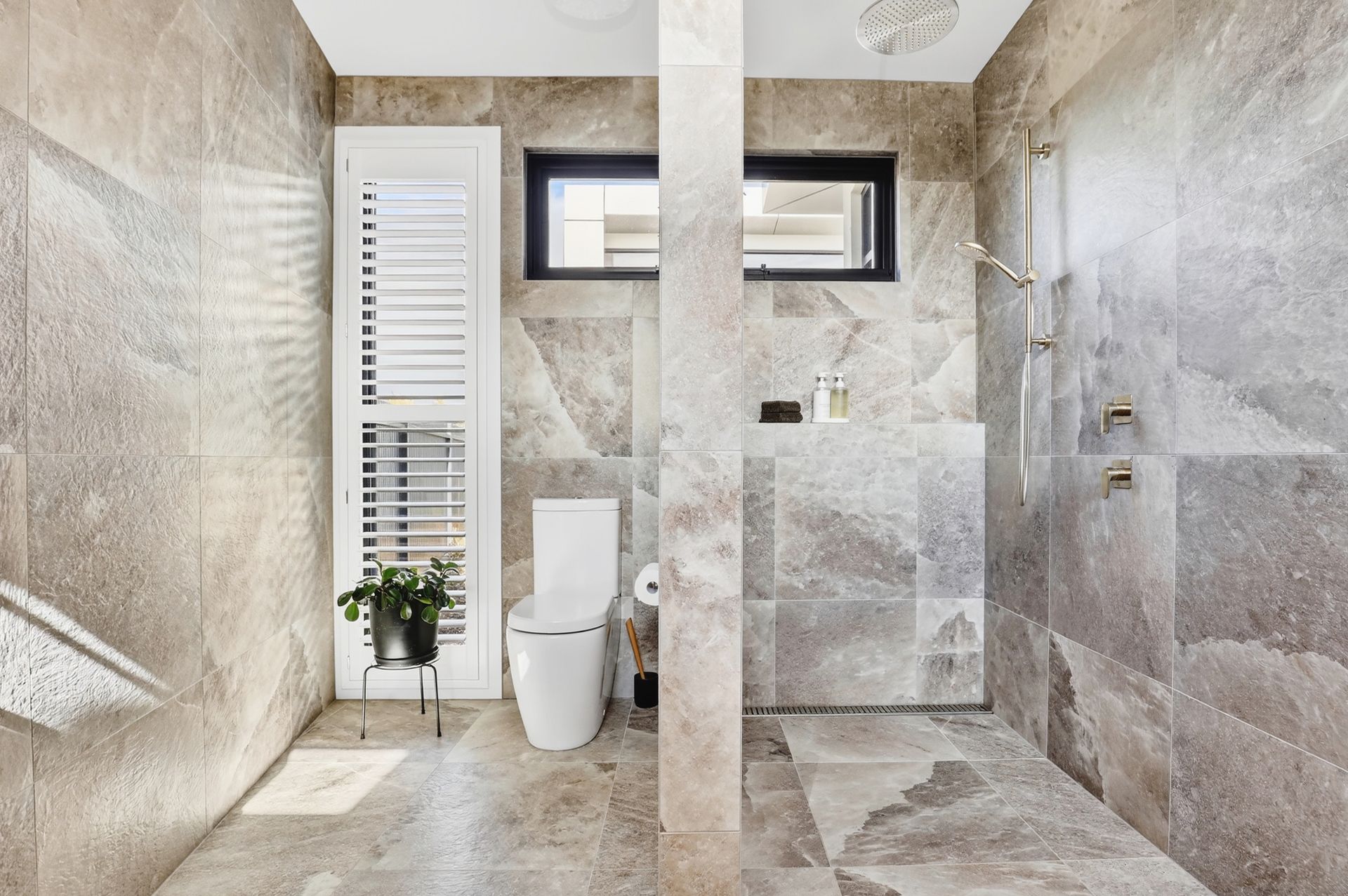 A modern bathroom with beige stone walls, toilet, shower, and white shutter door.