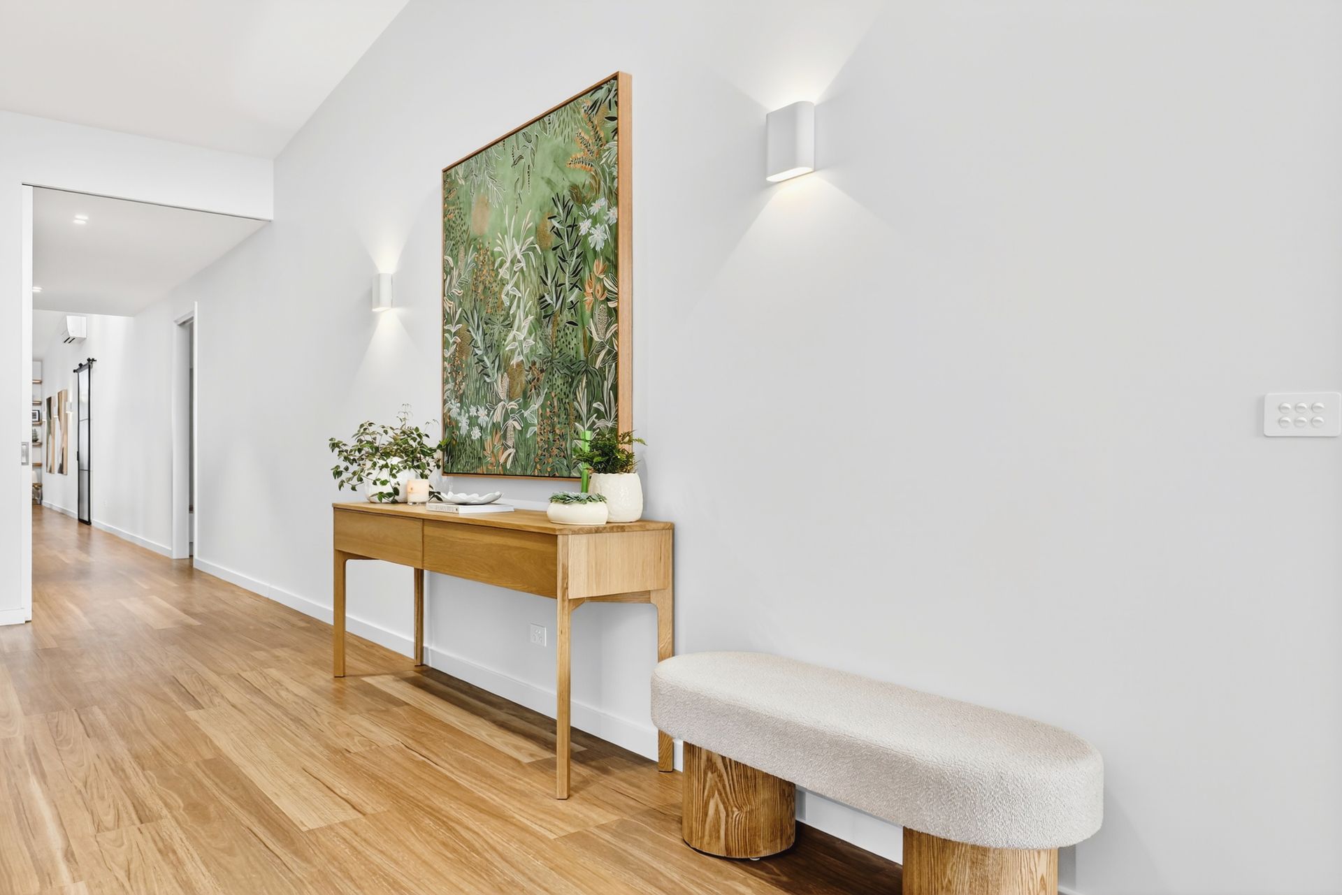 Hallway with wooden floor, console table, bench, artwork, and wall sconces.