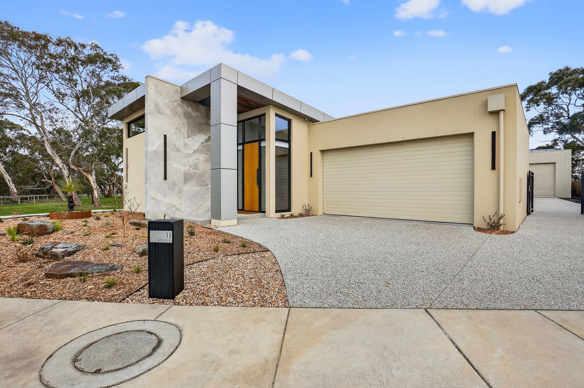 Modern beige house with gravel driveway, garage, and front yard landscaping under a blue sky.