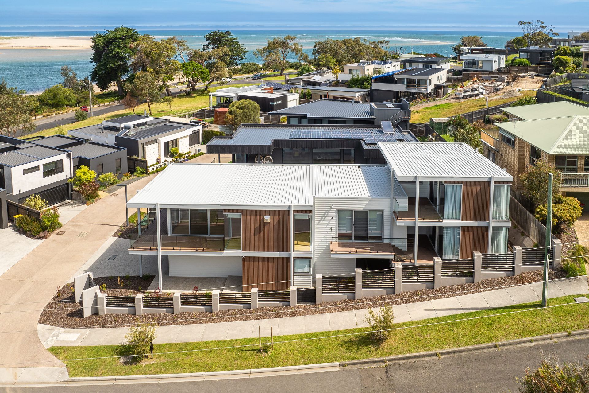Modern two-story house with a white roof and wooden accents, overlooking a coastal landscape with houses and water.