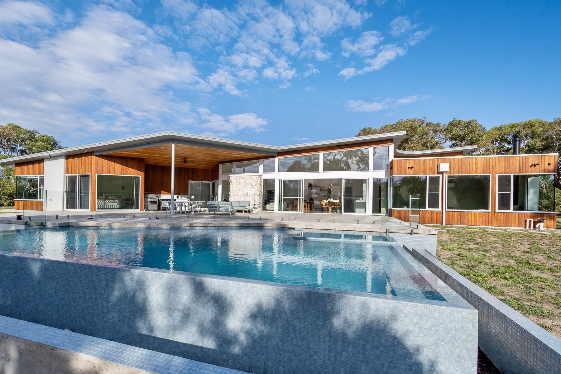 Modern home with a rectangular pool reflecting the blue sky and clouds. The house features wood paneling, large windows, and an open porch.