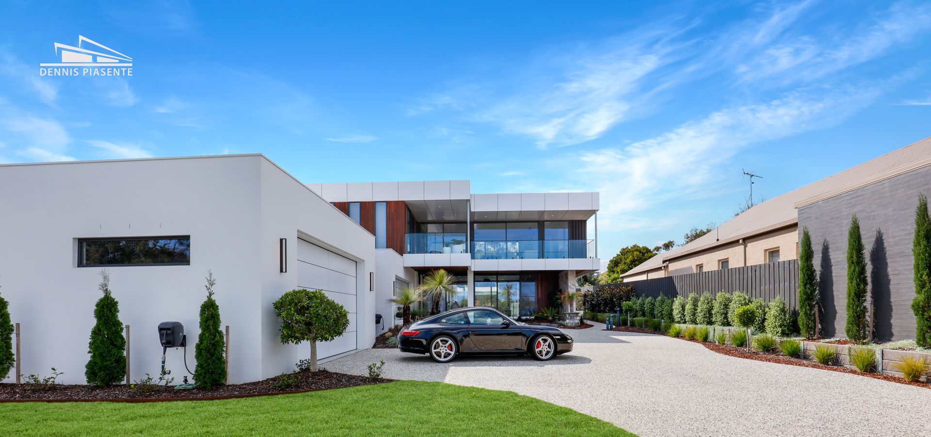 Modern house with a black sports car parked in the gravel driveway. The bright blue sky has wispy clouds.