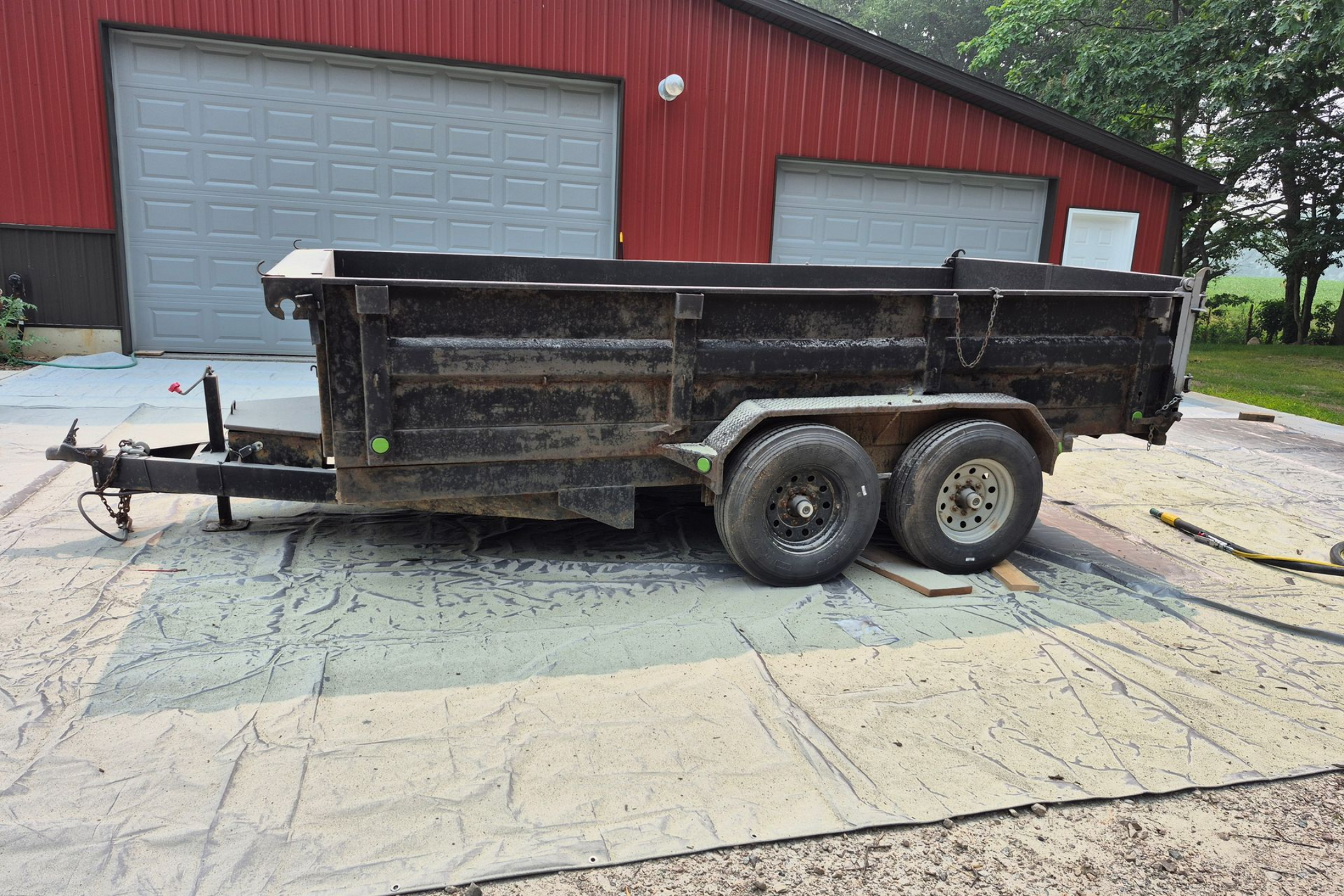 Black dump trailer parked on pavement in front of a red barn with gray garage doors