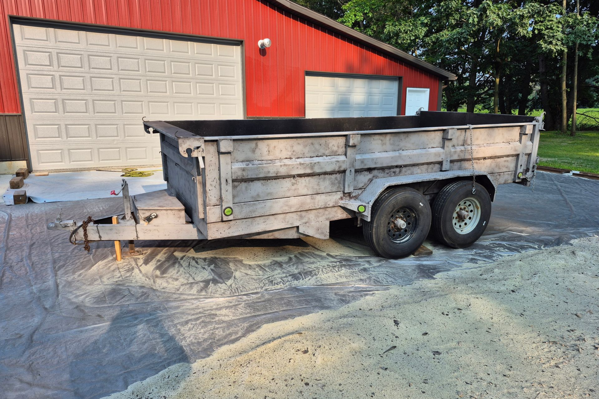 Utility trailer parked beside a red garage, with weathered wooden sides and dual axles.