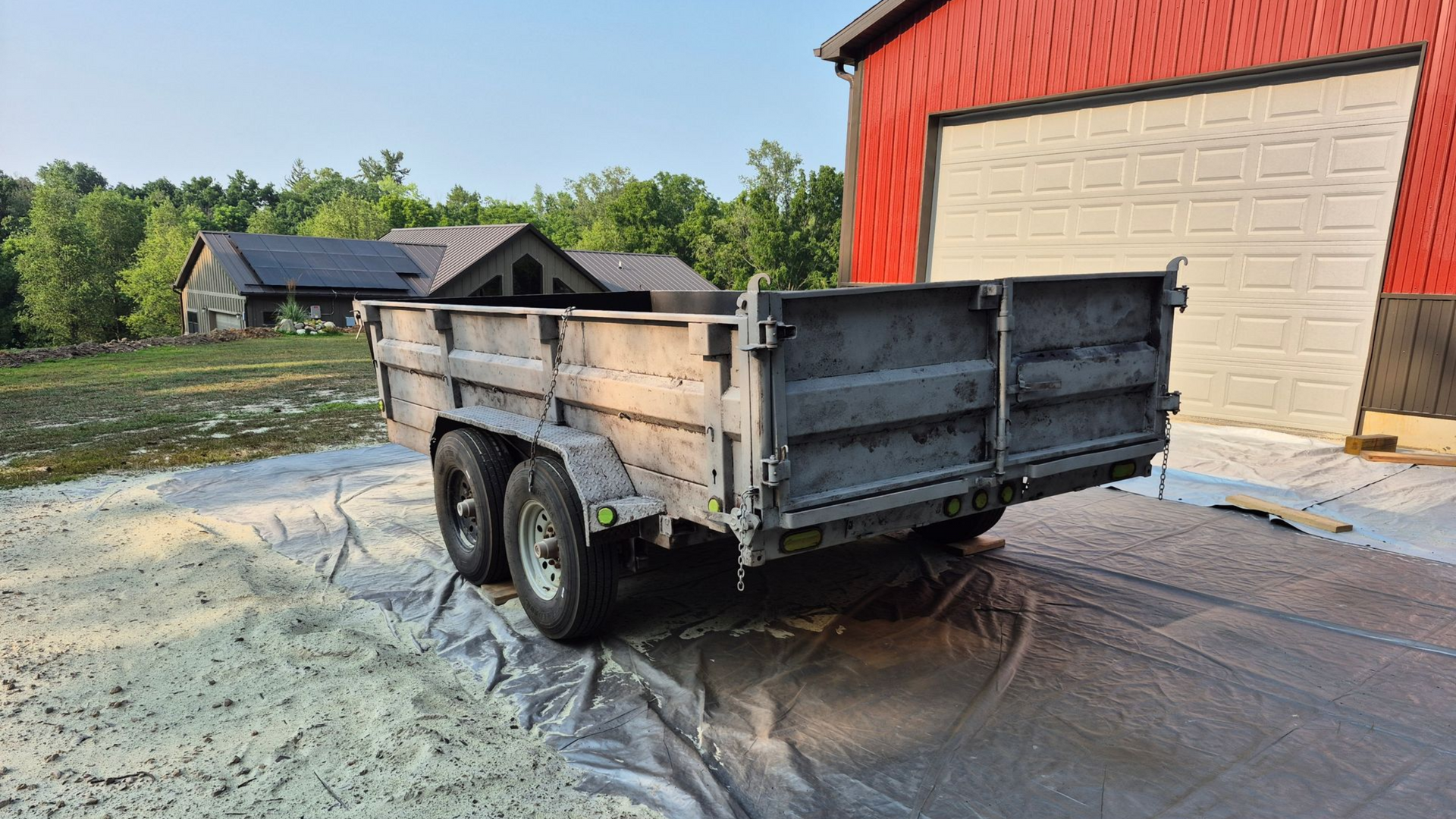 Small black utility trailer parked on a tarp beside a red garage on a sunny driveway