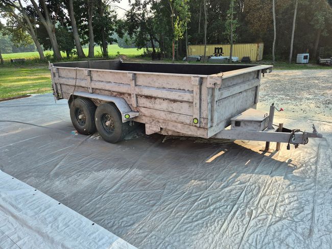 Metal dump trailer with dual wheels and hitch parked on a paved lot, with trees in the background