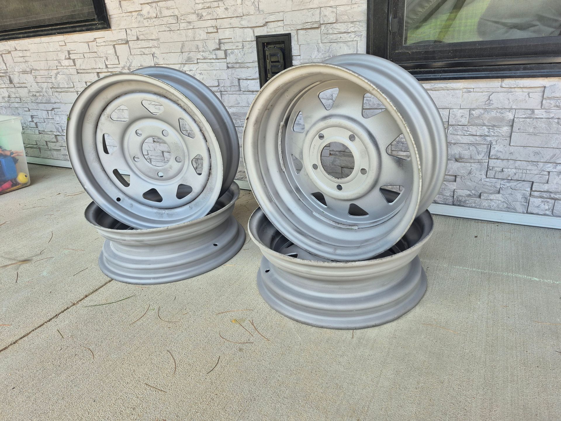 Two stacks of gray metal car rims leaning against a stone wall on a concrete driveway