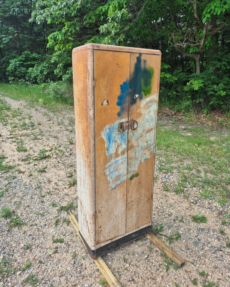 Weathered wooden cabinet or box outdoors on grass and dirt, with a faded blue door and foliage behind it