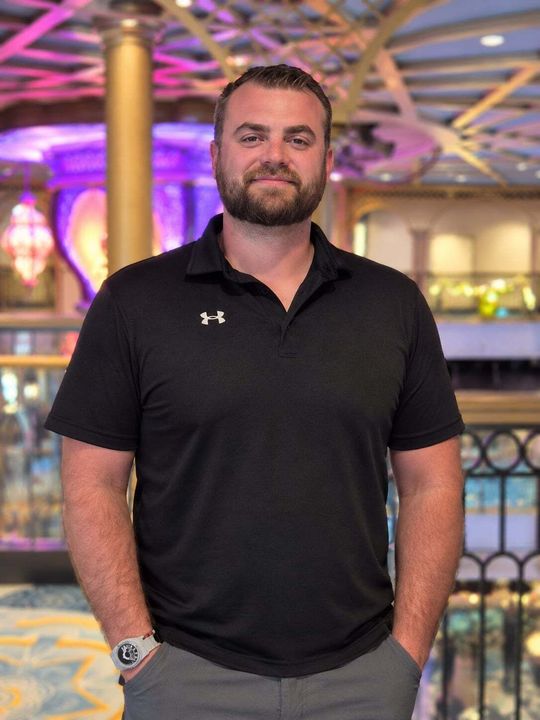 Man standing indoors in a black polo shirt with hands in pockets, lit by colorful ceiling lights