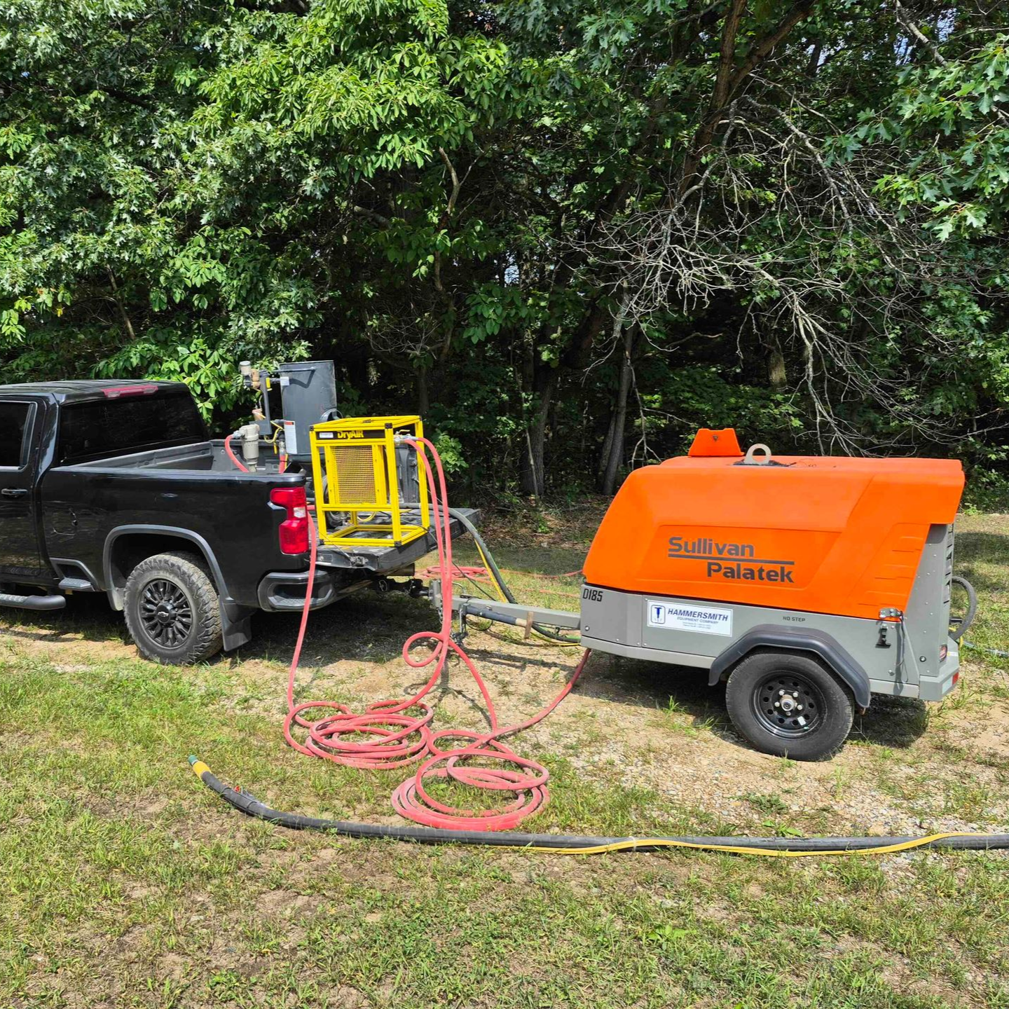 Black pickup truck and orange utility trailer with hoses and equipment in a grassy yard beneath trees