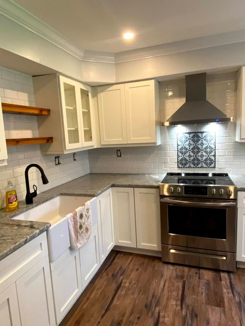 Bright white kitchen with gray countertops, wood floor, open shelves, and stainless steel stove under hood.
