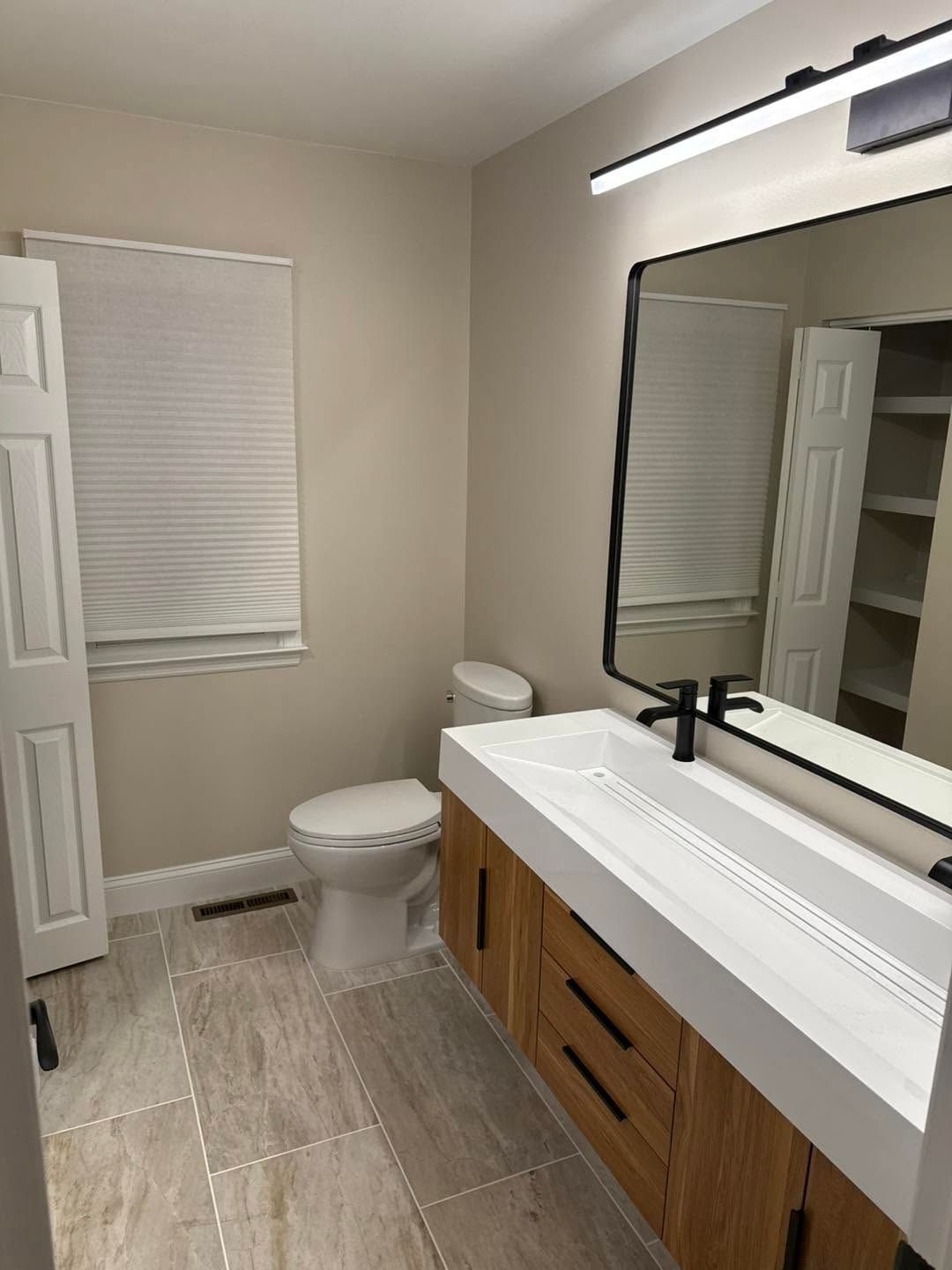Modern bathroom with double vanity, large mirror, toilet, and window with blinds