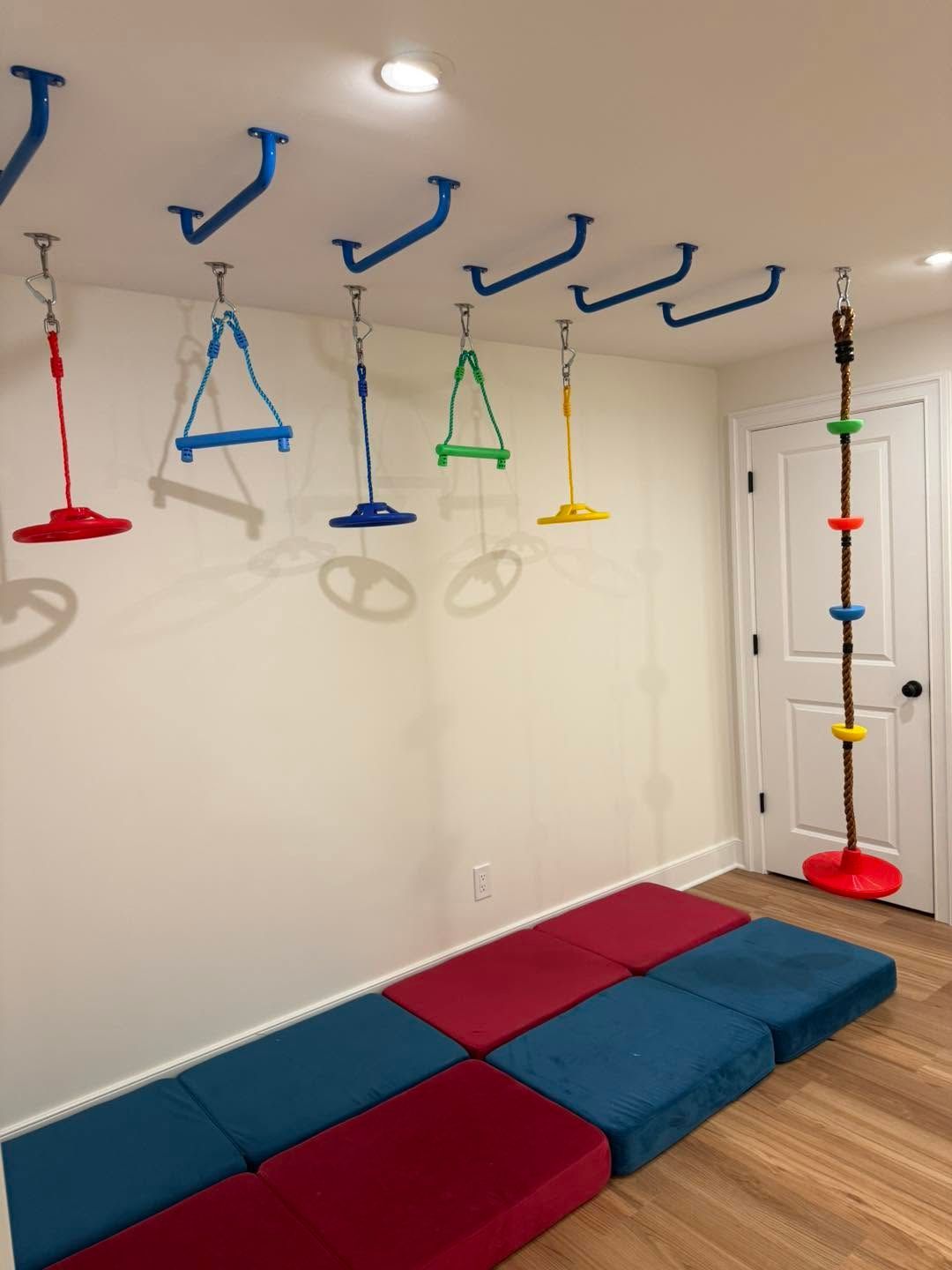 Indoor sensory gym with hanging swings, wall hooks, a climbing rope, and red and blue floor mats.