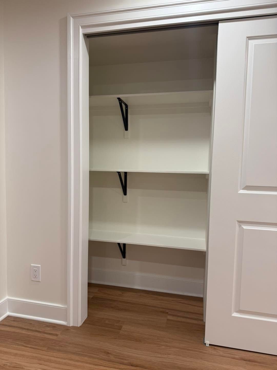 Open closet with white shelves and black brackets, in a room with hardwood floors and a sliding white door.
