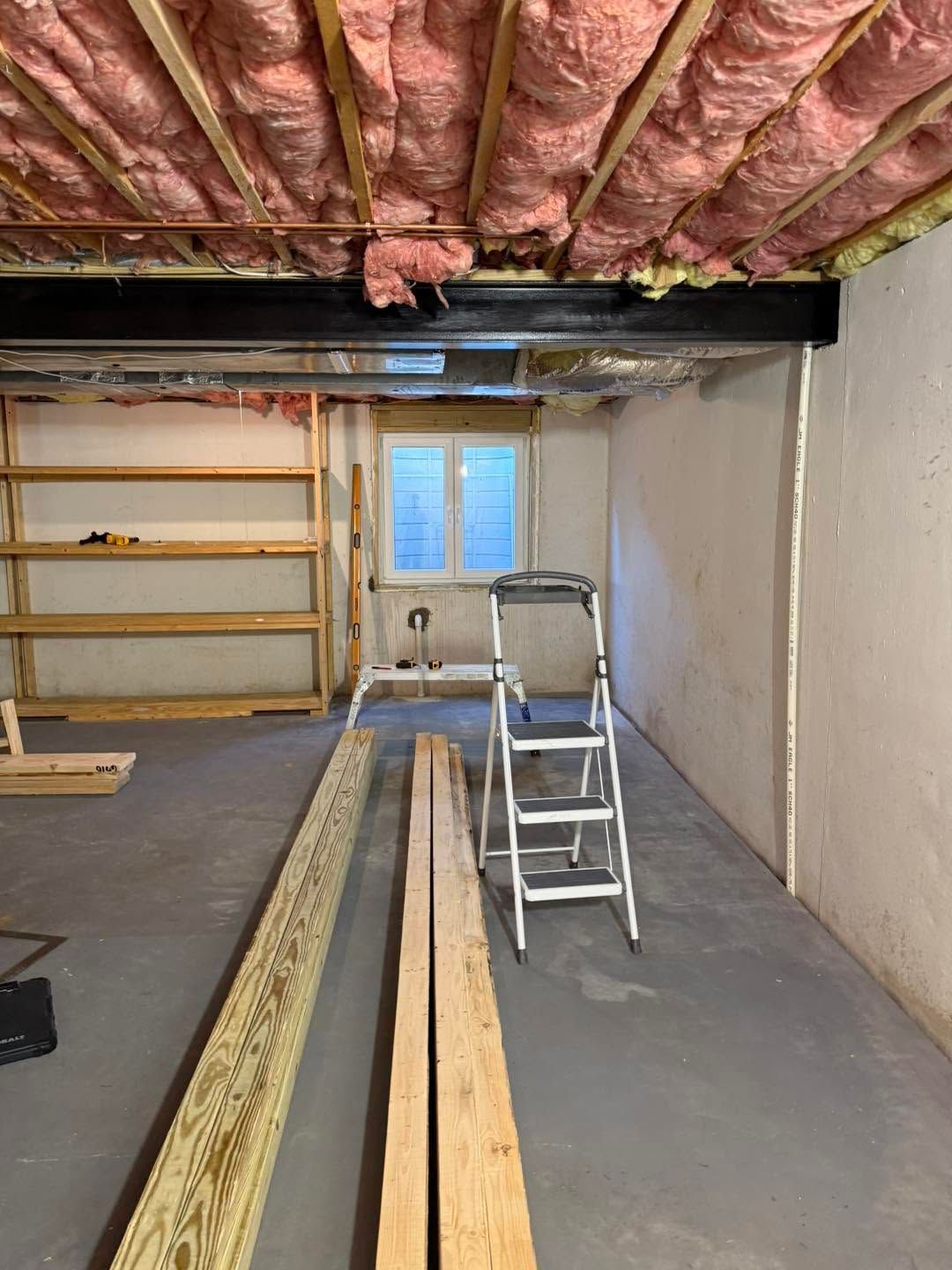Basement under renovation with exposed pink insulation, ladder, shelves, and lumber on the floor