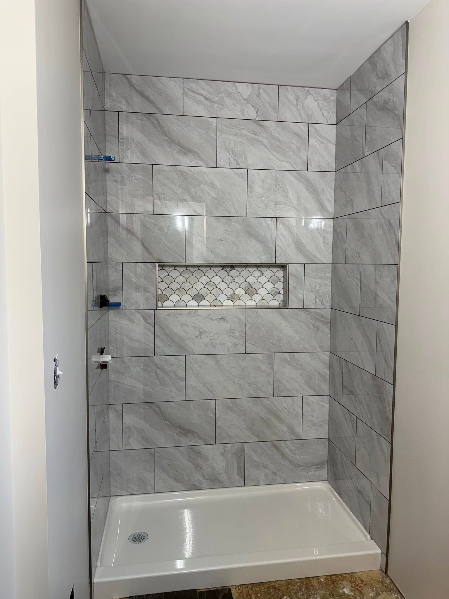 Tiled shower stall with white tub, gray marble-look walls, and built-in shelf niche
