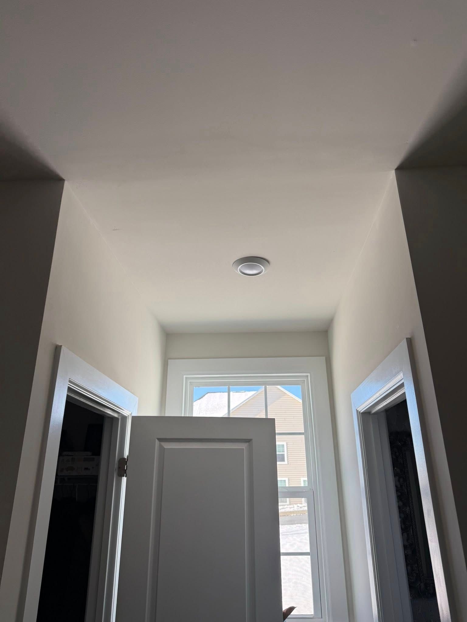 Hallway with white walls, doorways, and a window at the far end under a ceiling light.