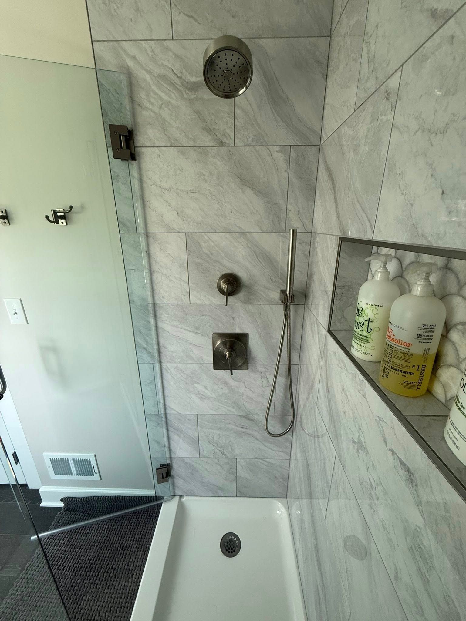 Bathroom shower with tiled walls, glass door, shelf of toiletries, and a white tub edge in the foreground