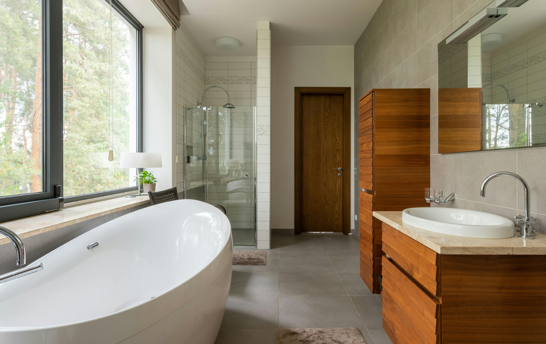 Modern bathroom with freestanding tub, glass shower, wood vanity, and large window with greenery outside