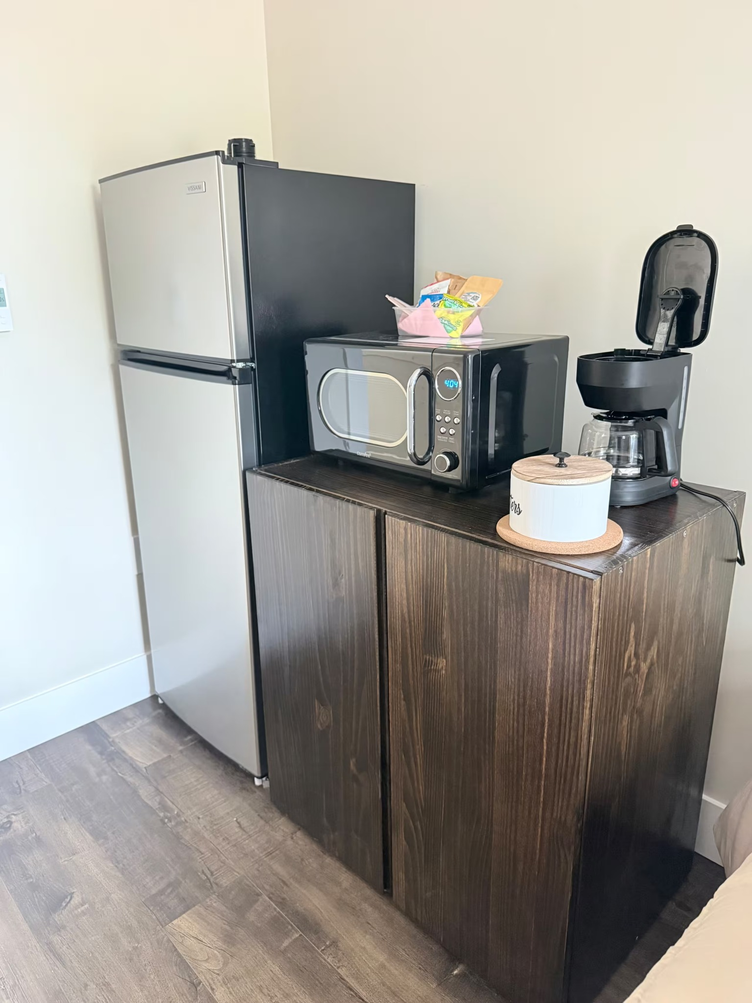 Compact kitchen corner with a stainless fridge, microwave, coffee maker, and small counter.