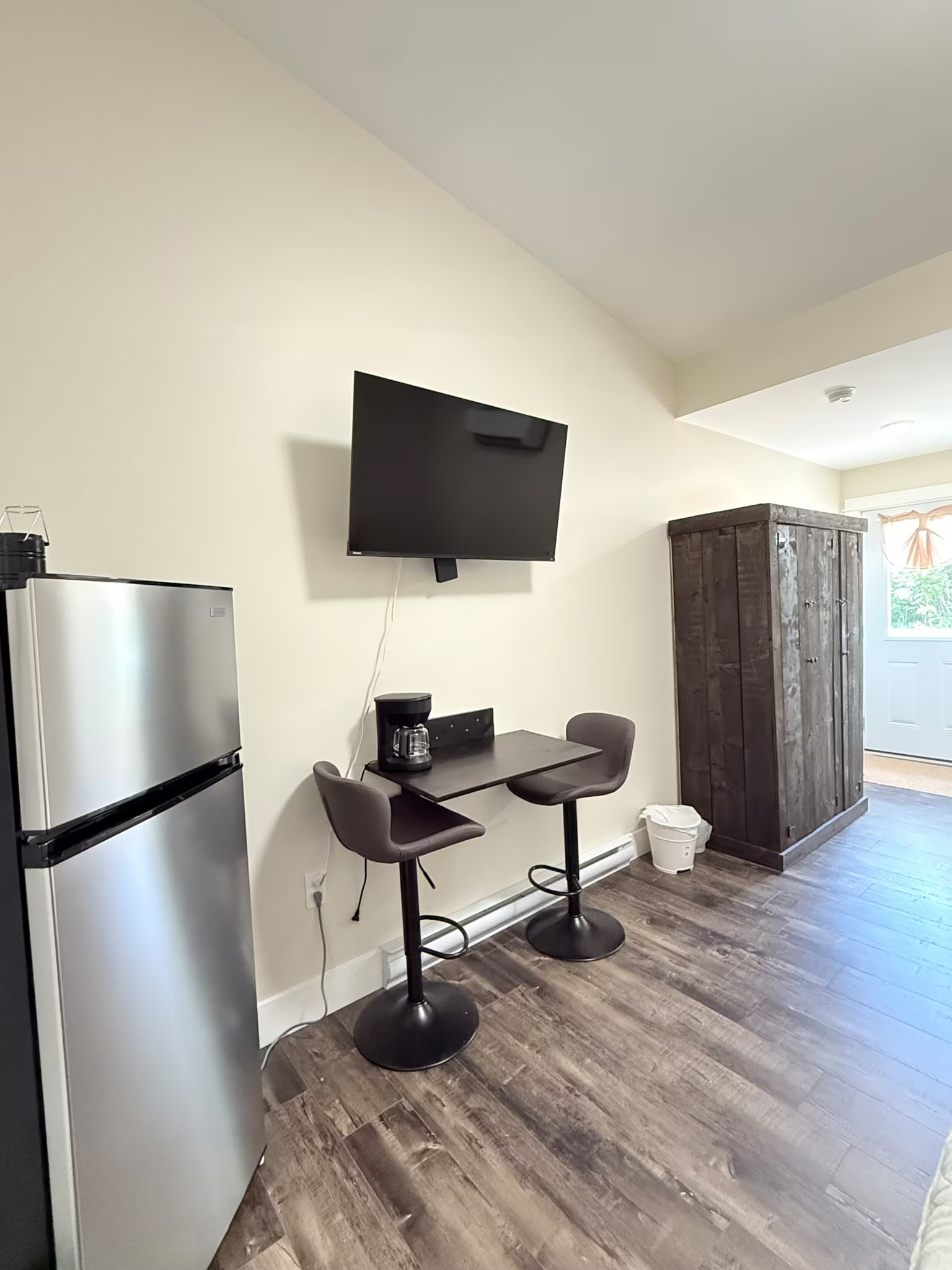 Small kitchenette with fridge, wall-mounted TV, two bar stools, and a tall cabinet on wood flooring.