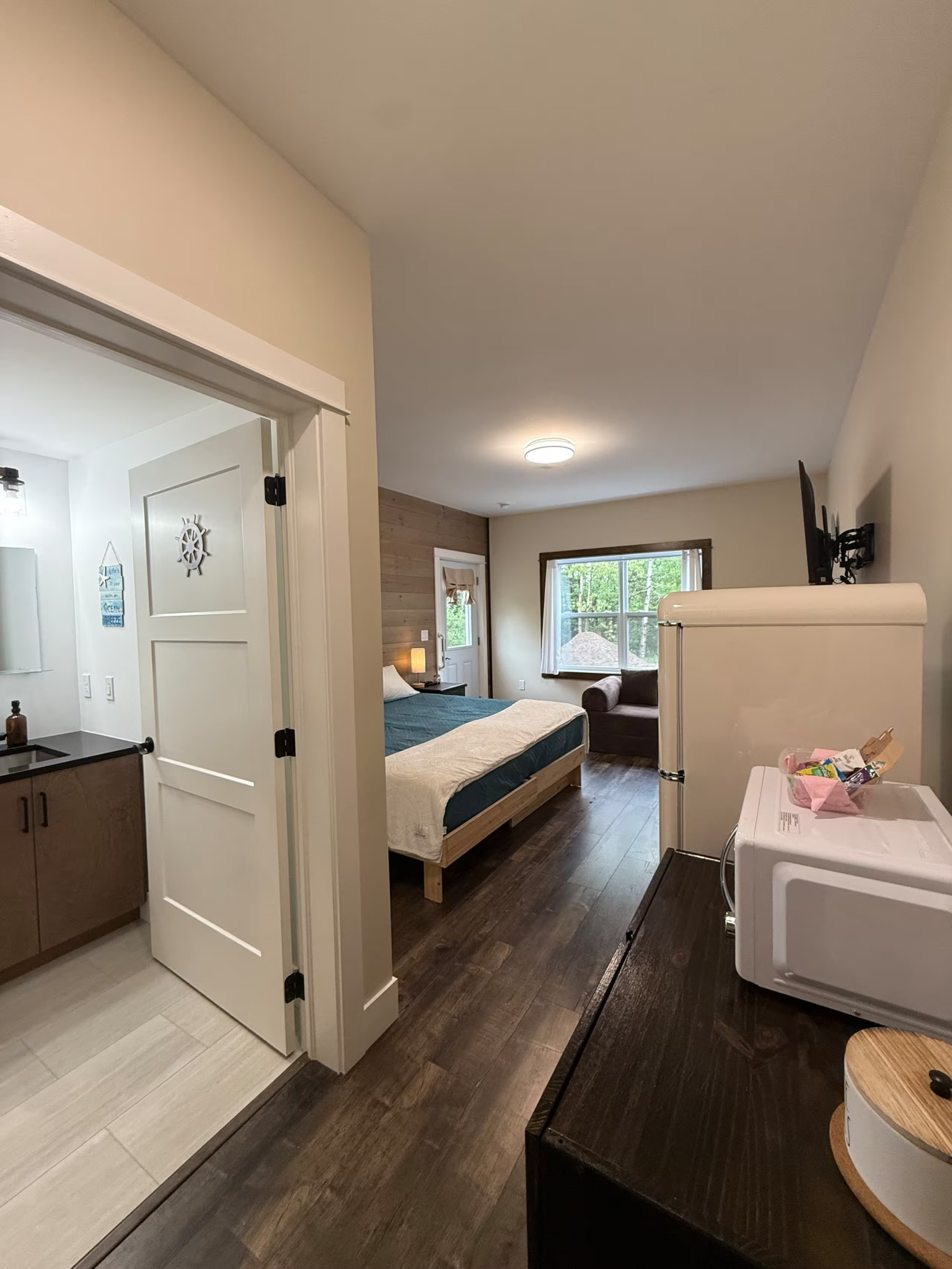 Bright bedroom with open bathroom door, white bed, dark wood floor, and large window overlooking greenery