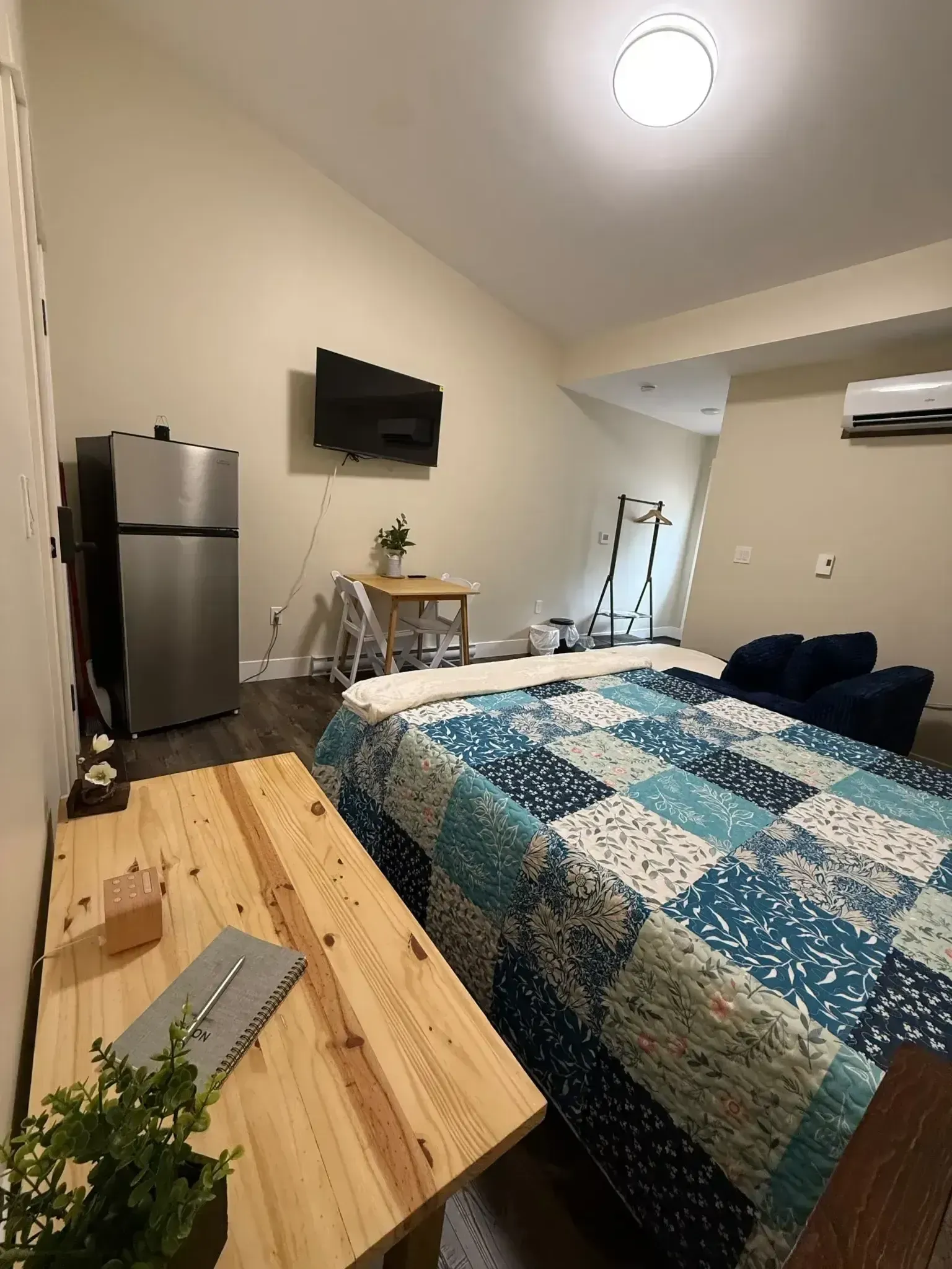 Small studio bedroom with bed, fridge, wall-mounted TV, desk, and hardwood floor; blue quilt on bed.