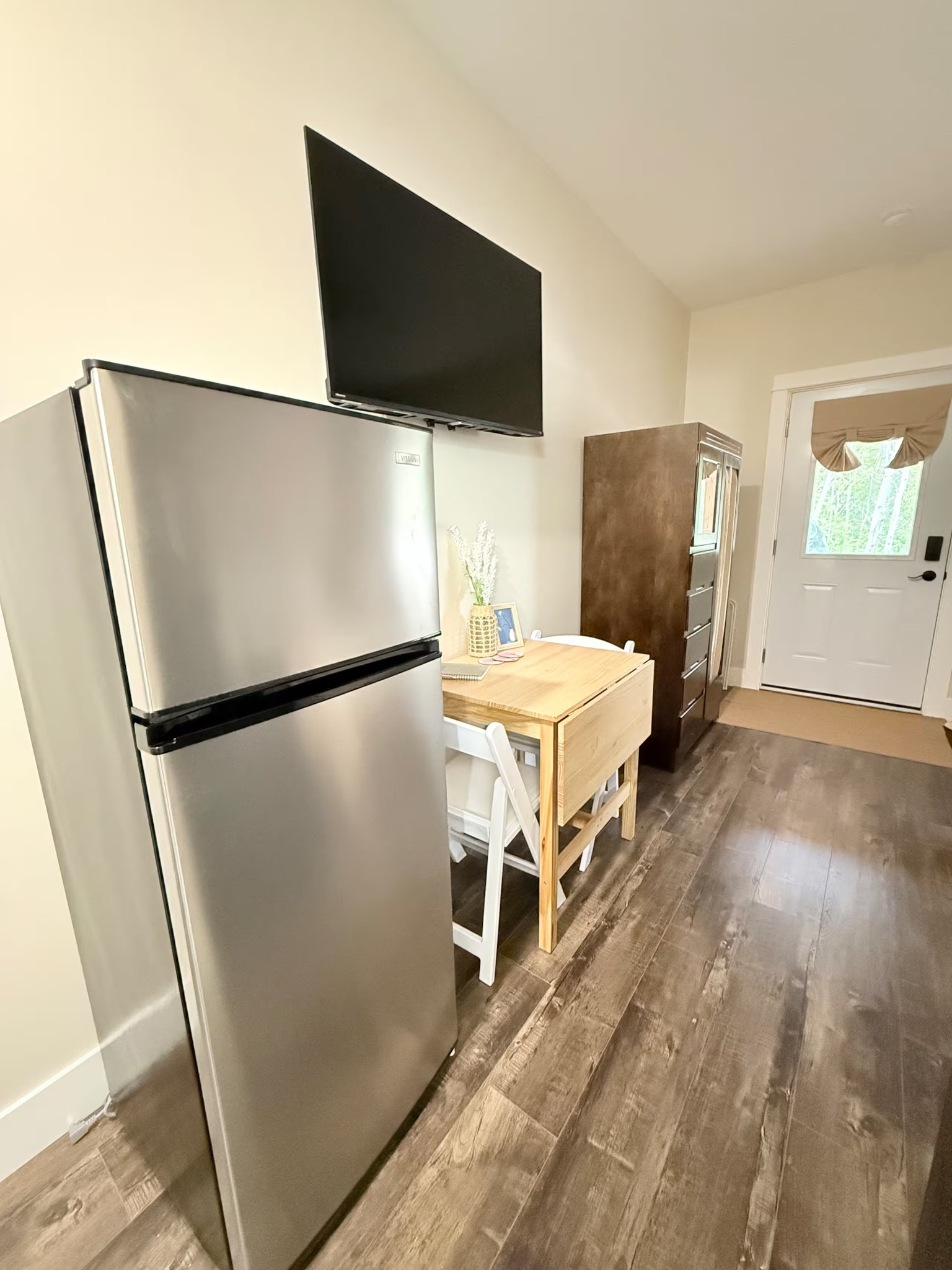 Small kitchen with a stainless refrigerator, TV on the wall, and a table by a doorway.