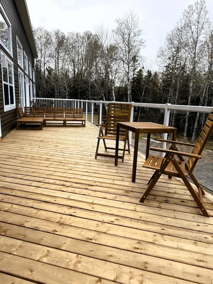 Wooden deck with outdoor table, chairs, and bench beside a house, overlooking leafless trees.