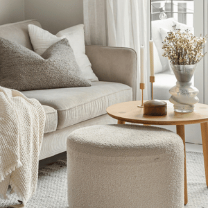 Cozy beige living room with a sofa, ottoman, round coffee table, and vase of dried flowers.