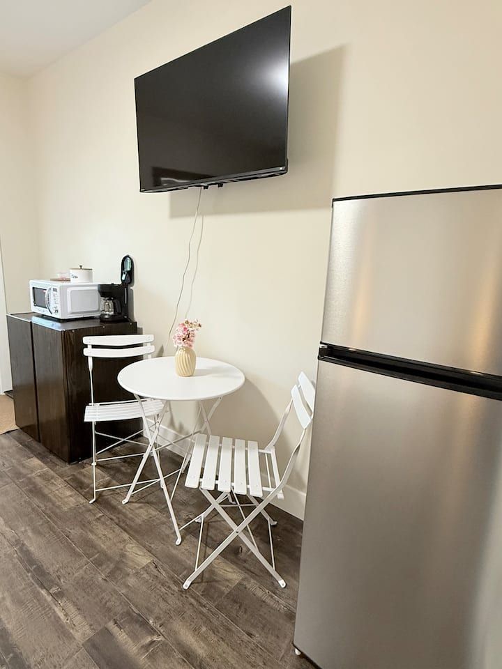 Small kitchenette with wall-mounted TV, stainless fridge, coffee maker, and a round table with folding chairs.