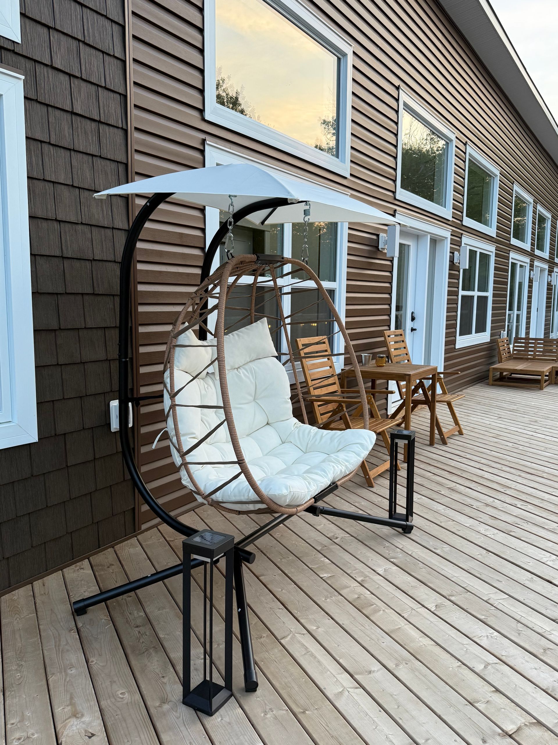 White hanging egg chair with blue cushion on a wooden deck outside a building with windows