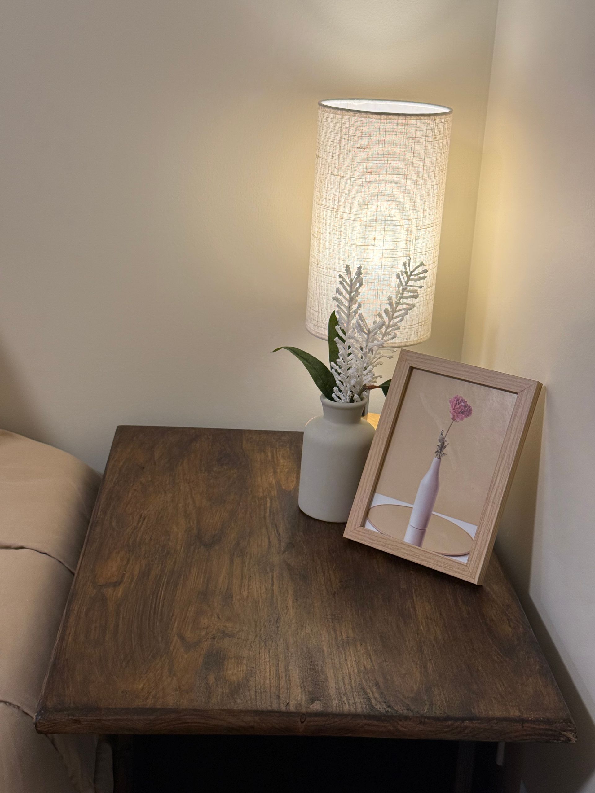 Wooden table with a lit white lamp, small vase, and framed floral art against a beige wall.