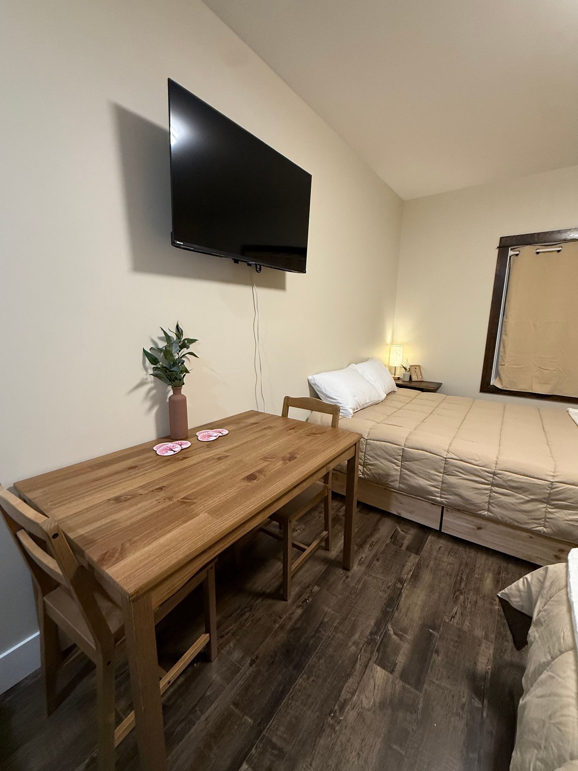 Small bedroom with wall-mounted TV, wooden table, bed, and nightstand lamp