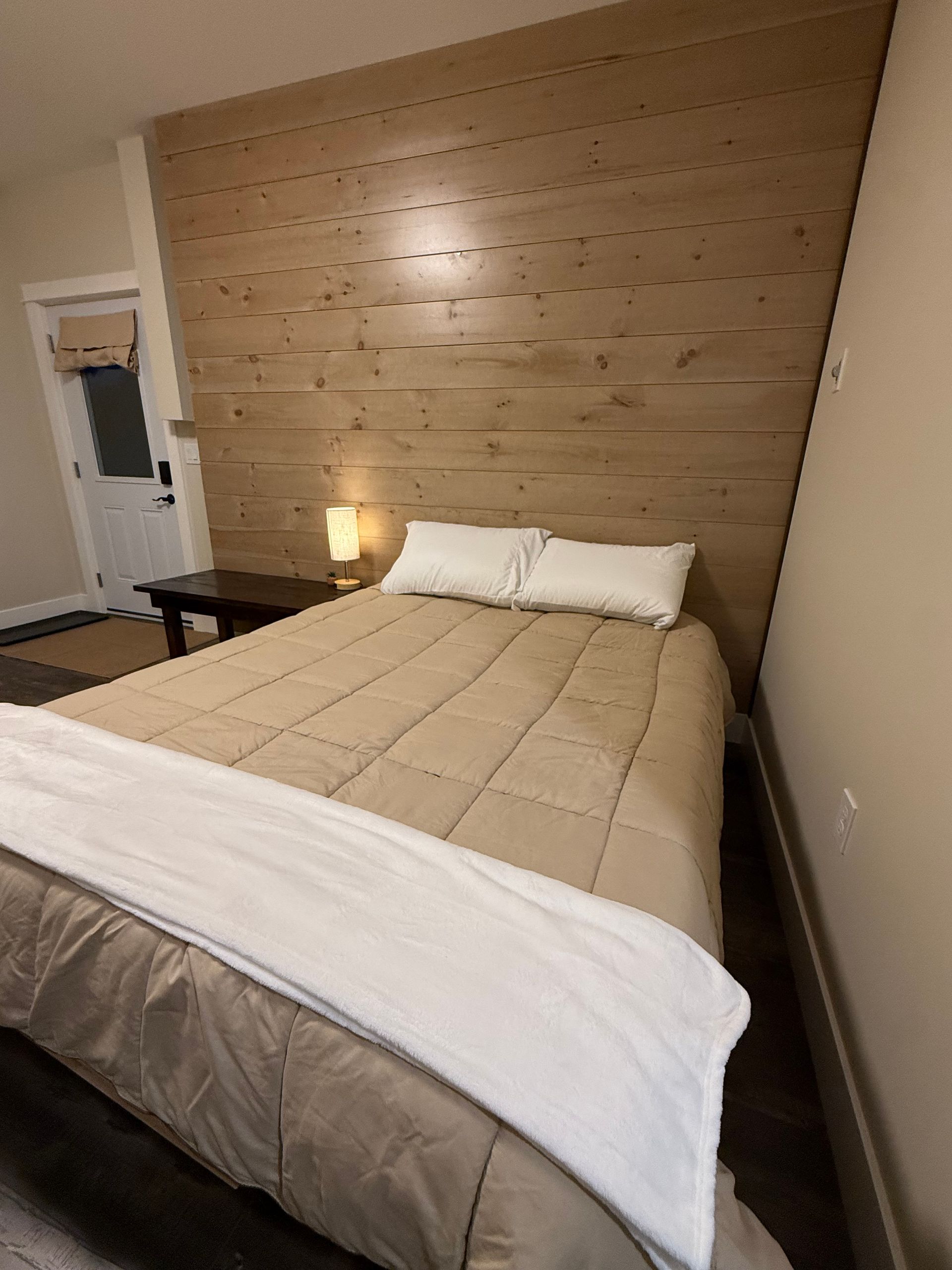 Bedroom with a neatly made bed, tan quilt, white pillows, and a wood-paneled accent wall.