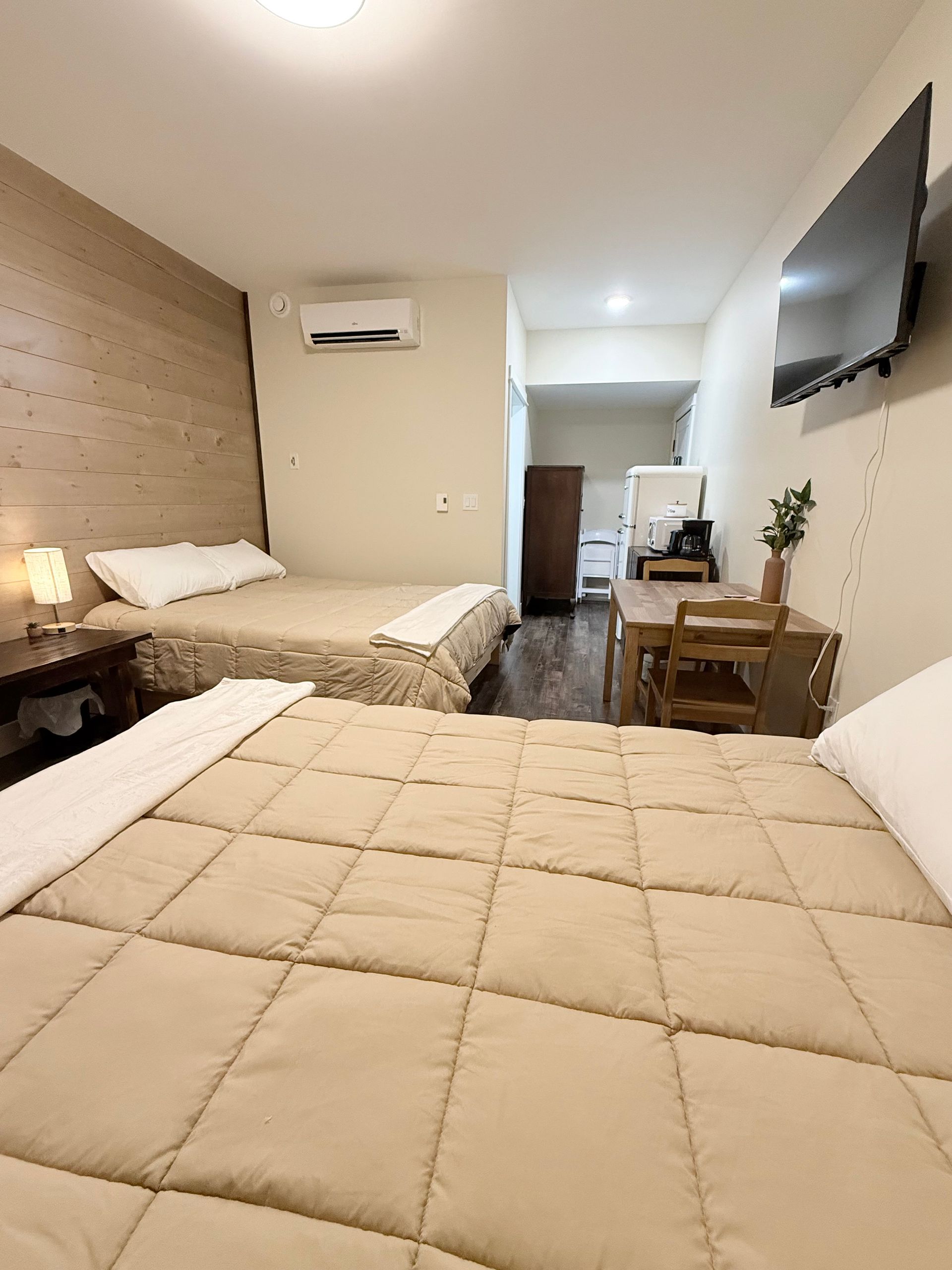 Cozy hotel room with two beds, wood accent wall, wall-mounted TV, and a small desk near the doorway